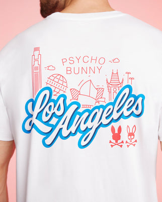 A man wearing a retro Dodgers tee showcases his style with "Los Angeles" boldly written in blue and white script on a white Pima cotton shirt. Iconic red outlines of LA landmarks sit above the text along with "Psycho Bunny," while two pink bunny logos adorn the bottom. The shirt is the MENS LOS ANGELES CITY TEE - B6U861A2PC by Psycho Bunny.