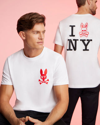 Two men wearing matching Pima cotton jersey white t-shirts with a red bunny and crossbones logo. The man in the foreground faces forward, while the man in the background faces away, revealing text on his Psycho Bunny MENS NEW YORK TEE - B6U554W1PC that reads "I [bunny with crossbones] NY." The background is pink.