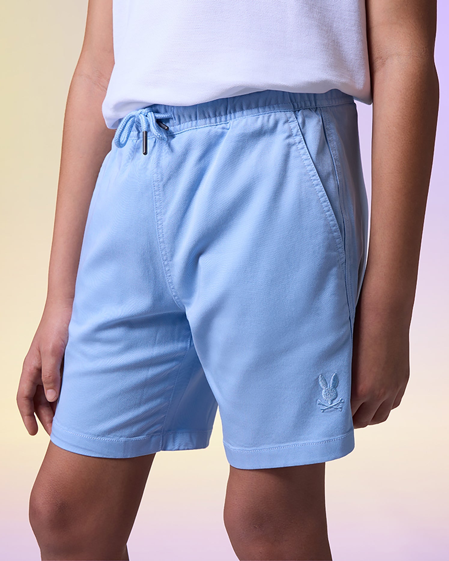 A person wearing light blue drawstring shorts made from Tencel-blend twill and a white top stands in front of a gradient background ranging from light yellow to light purple. The KIDS WILLIS STRETCH TENCEL SHORT - B0R239Y1WB by Psycho Bunny, featuring pockets and an embroidered logo near the bottom hem, epitomize elevated ease for your summer go-to outfit.