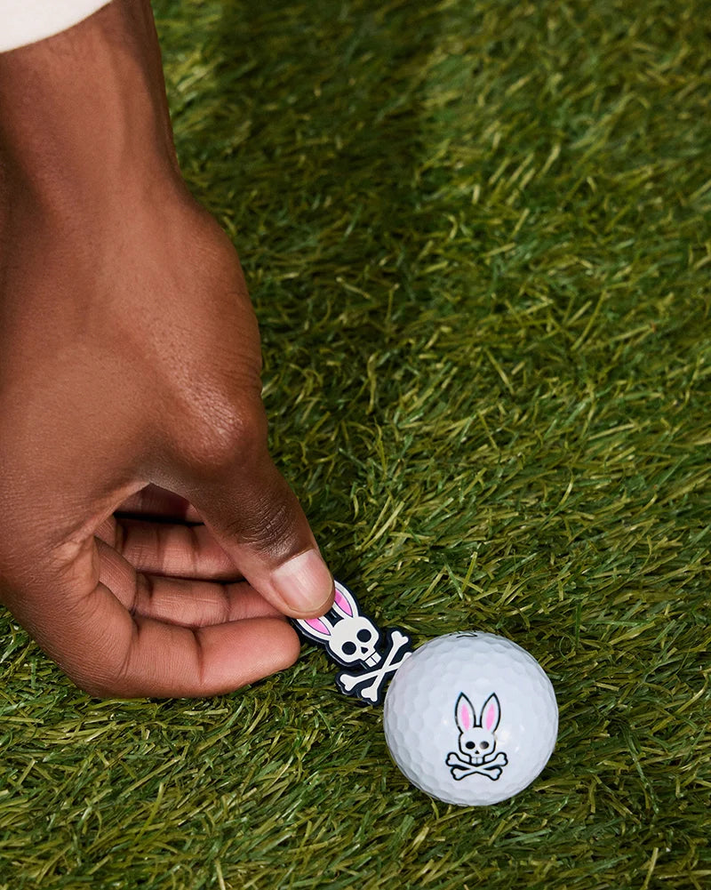 A close-up image of a person's hand holding the BALL MARKER - B6A698C200 by Psycho Bunny, showcasing its distinct skull with bunny ears design. The marker is placed next to a golf ball featuring the same edgy aesthetic. Both items, part of the exclusive golfers collection, rest harmoniously on grassy turf.