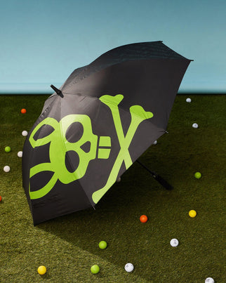 A black Psycho Bunny GOLF UMBRELLA - B6A624C200 featuring a large green skull and crossbones design lies open on a grass-like surface. Surrounding the umbrella are colorful golf balls scattered around, adding a playful element for die-hard golfers. The background is a light blue gradient.