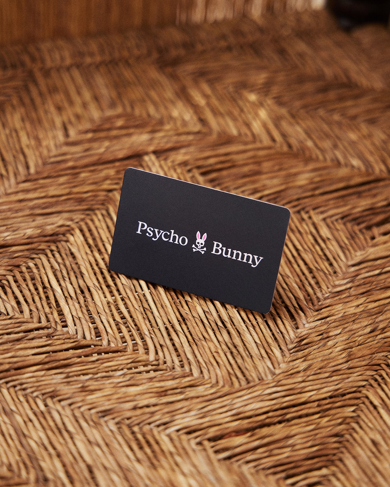 A black rectangular Psycho Bunny Canada Digital Gift Card rests on a textured, woven surface. The card features the logo and text 