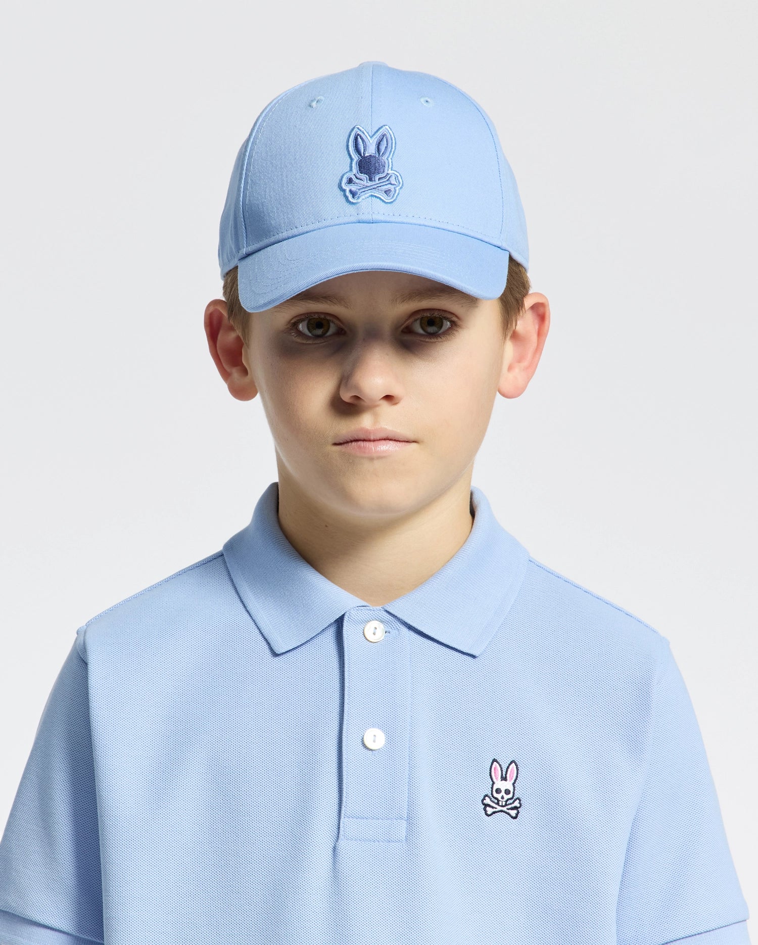 A young boy wears a light blue cotton polo with a rabbit emblem and the Psycho Bunny KIDS NEW CASTLE EMBROIDERED BASEBALL CAP - B0A547F200. The cap has an adjustable back strap for a perfect fit. He stands against a plain white background, gazing neutrally at the camera.