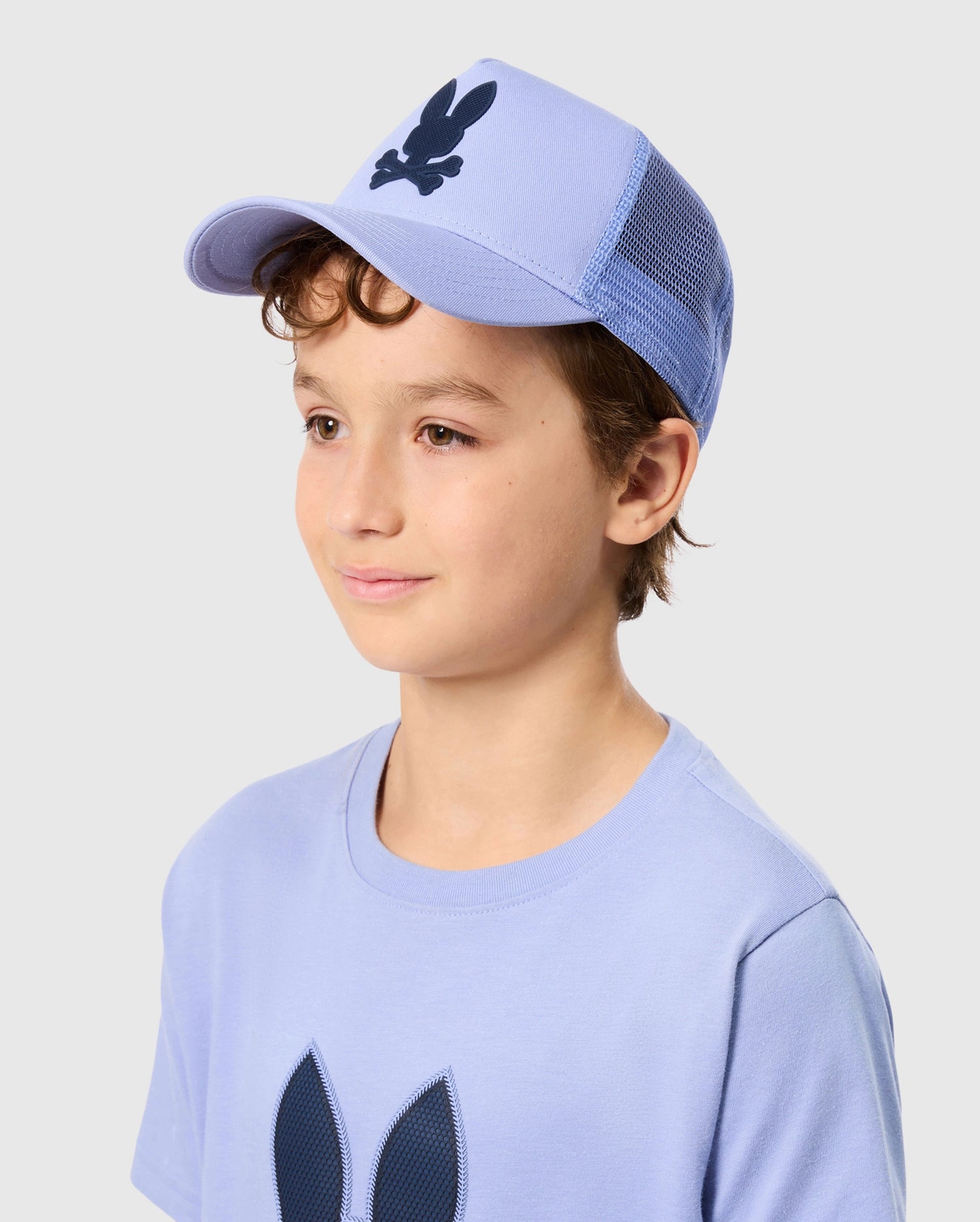 A young boy with curly hair is wearing a light blue KIDS RIVIERA TRUCKER CAP by Psycho Bunny and a matching T-shirt featuring the brand's dark blue bunny logo. He is looking to the side with a neutral expression, evoking early 2000s nostalgia against the plain light gray background.