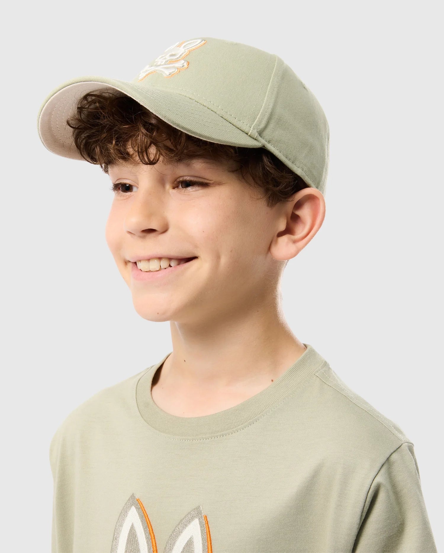 A young boy with curly brown hair is smiling and looking to the side. He is wearing a light green Psycho Bunny KIDS KAYDEN BASEBALL CAP - B0A677C200 and a matching t-shirt featuring a Bunny logo appliqué. The background is plain light gray.