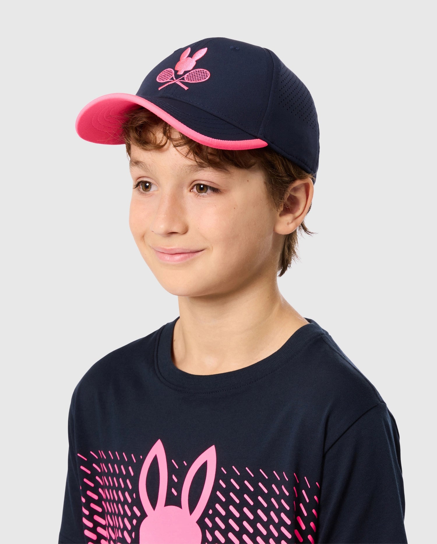 A child wearing a navy blue KIDS TORONTO TOURNAMENT CAP - B0A704C200 by Psycho Bunny, which features an embroidered tennis bunny design, along with a matching navy blue t-shirt showcasing a pink bunny graphic. The child has short brown hair and is smiling softly, with a grey background behind them.