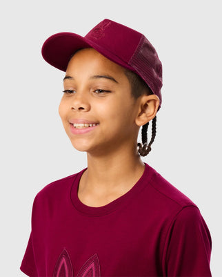 A young person with braided hair is smiling, wearing a stylish KIDS MALTON TRUCKER CAP - B0A797D200 by Psycho Bunny and a matching T-shirt with a graphic design on the front. The background is plain light gray.