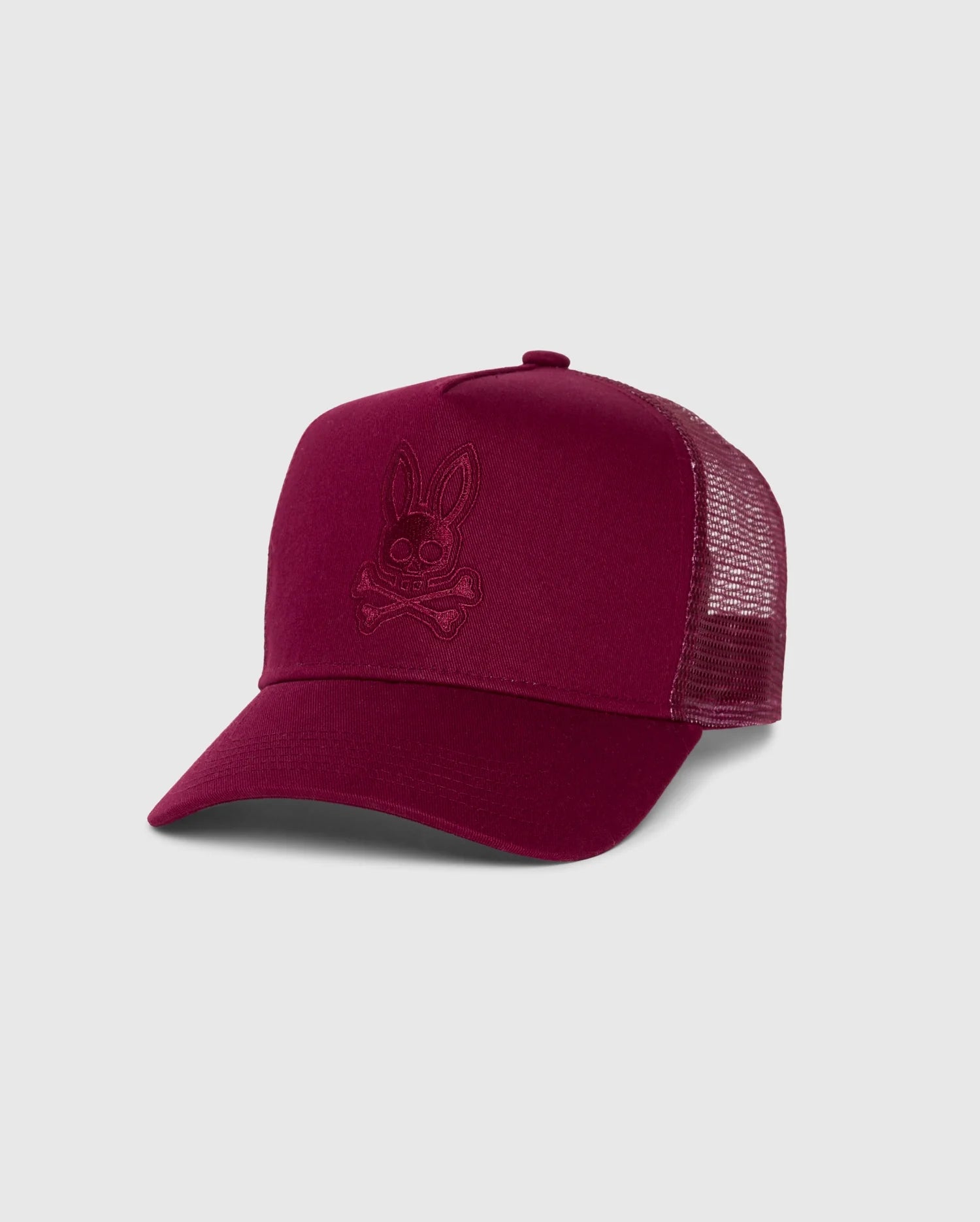 The Psycho Bunny Kids Malton Trucker Cap (B0A797D200) is a stylish maroon cap featuring a tonal embroidered bunny skull and crossbones design on the front, complemented by a mesh back. The cotton baseball cap is set against a plain light gray background.