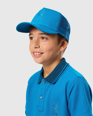 A young person with light skin and short brown hair is wearing a blue baseball cap, identified as the KIDS MALTON TRUCKER CAP - B0A797D200 from Psycho Bunny, along with a blue polo shirt. The cap, featuring mesh ventilation, and the shirt both display a small embroidered rabbit logo. Both items are set against a plain, light-colored background.