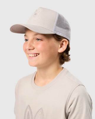 A young person wearing a beige Psycho Bunny Kids Malton Trucker Cap (B0A797D200) and a matching beige t-shirt is smiling and looking to the side. The cap, made from 100% cotton with mesh ventilation at the back, features an embroidered design on the front. The background is a plain light grey.