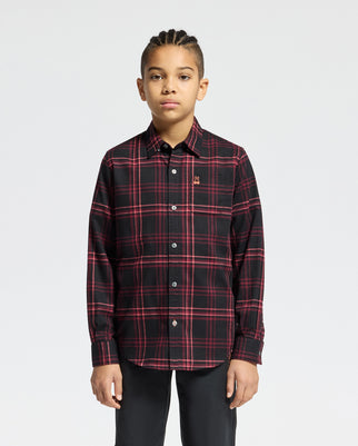 A young person with braided hair is stylishly dressed in the KIDS HAYES PLAID SHIRT - B0C842D200 by Psycho Bunny, made from soft Pima cotton, paired with black pants, standing against a plain white background.