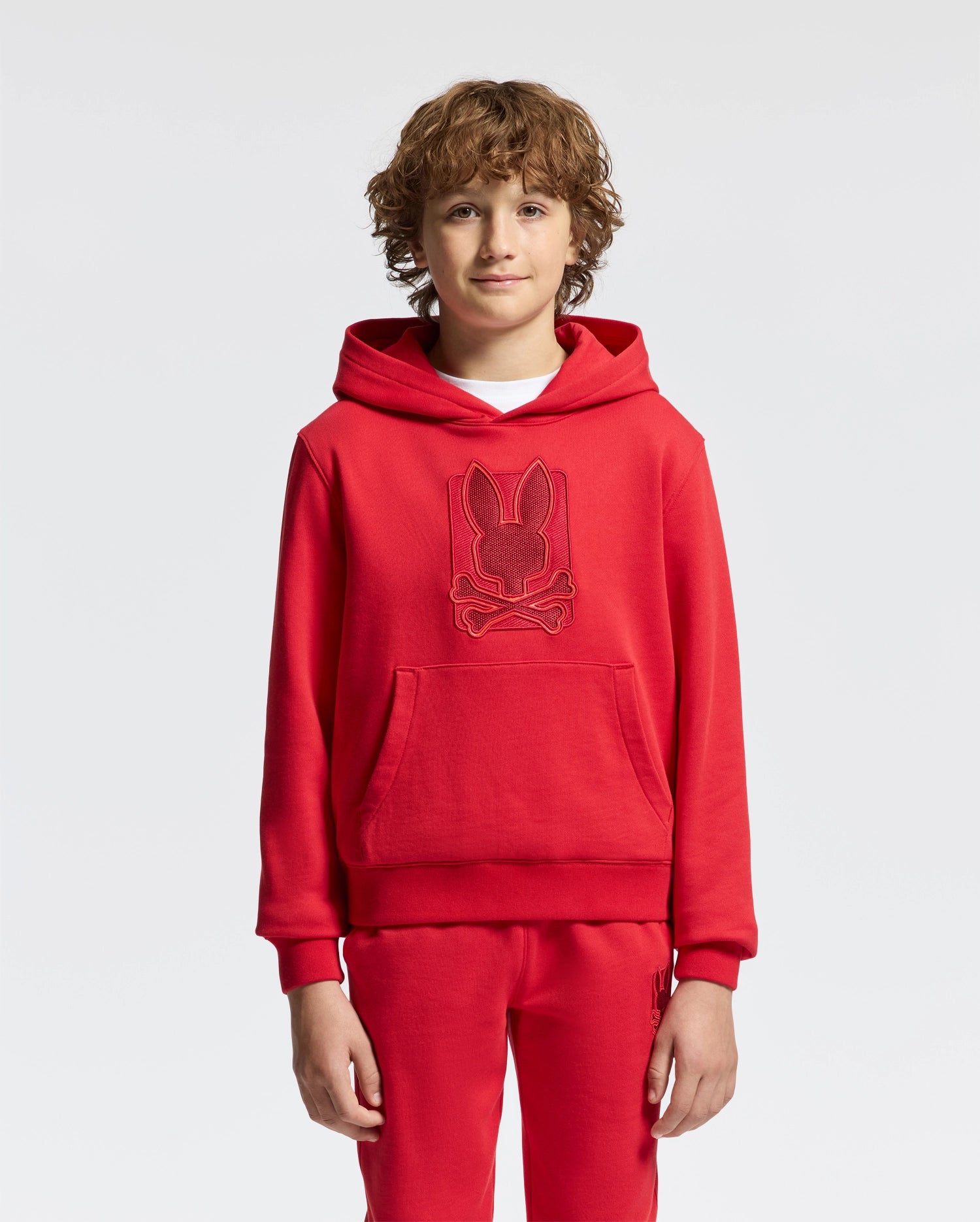 A young boy with curly hair stands wearing a vibrant red KIDS PIERCE ESSENTIAL FRENCH TERRY HOODIE from Psycho Bunny, along with matching pants. The hoodie, made of cotton, showcases an embroidered design of a rabbit's head with crossbones underneath it against a plain white background.