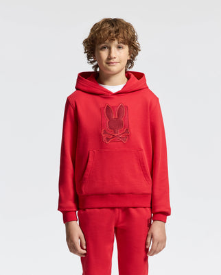 A young boy with curly hair stands wearing a vibrant red KIDS PIERCE ESSENTIAL FRENCH TERRY HOODIE from Psycho Bunny, along with matching pants. The hoodie, made of cotton, showcases an embroidered design of a rabbit's head with crossbones underneath it against a plain white background.