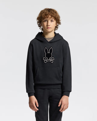 A young individual with curly hair is seen wearing a stylish Psycho Bunny Kids Alexander Hoodie (B0H154E200) in black, adorned with a bunny and crossbones motif on the front. The hoodie’s comfortable, relaxed fit enhances their fashionable appearance as they pose against a simple, light-colored background.