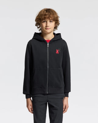 A young person with curly hair is wearing the Psycho Bunny KIDS PIERCE ESSENTIAL FRENCH TERRY ZIP HOODIE, paired with black pants, while facing the camera. The zip hoodie features a red graphic on the left side, adding flair to the look against the plain white background.