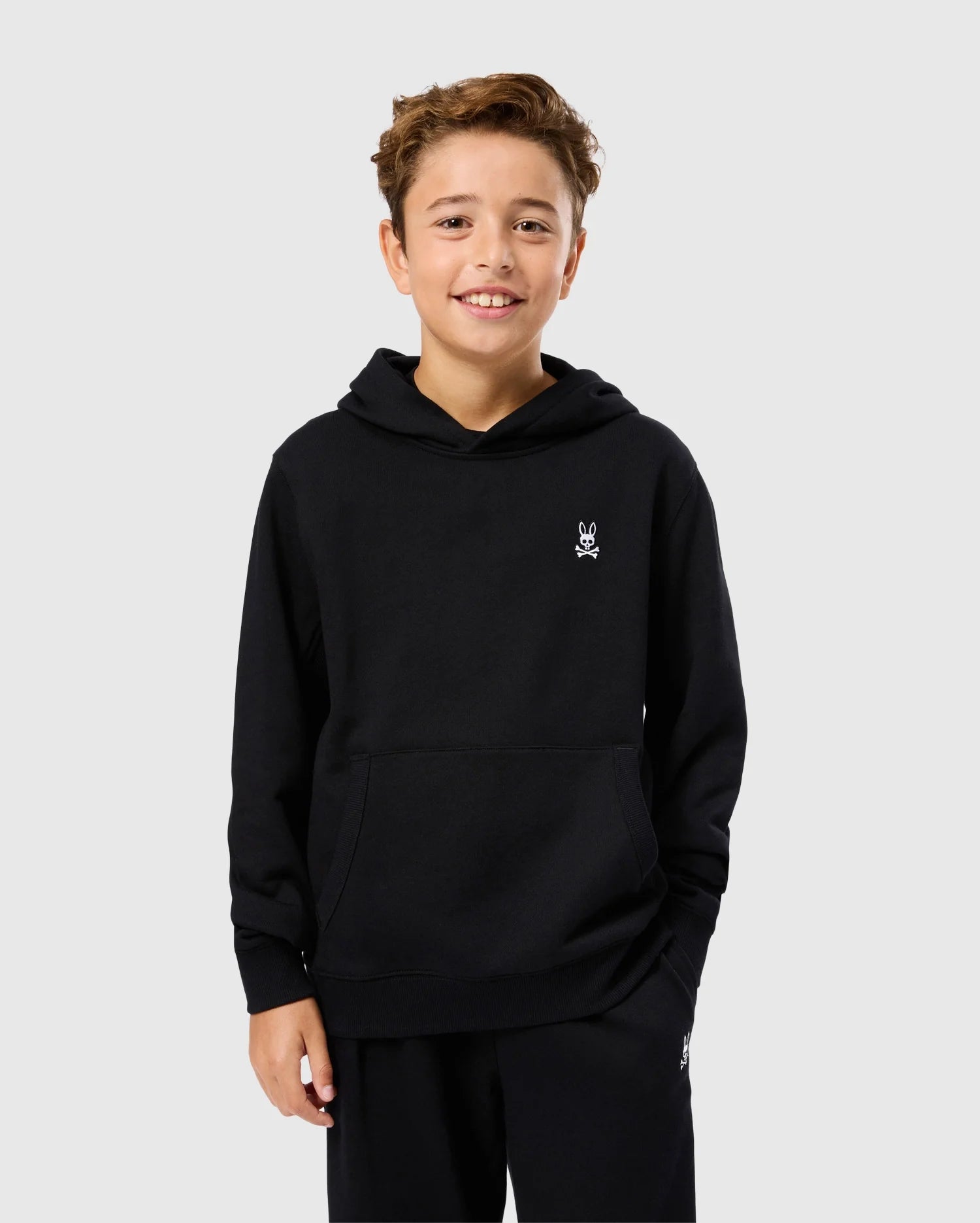 A smiling boy wears the Psycho Bunny KIDS LEVY FLEECE HOODIE (B0H859D200), featuring a regular fit in black micro-French terry with a small white logo on the chest. He stands against a plain light gray background.