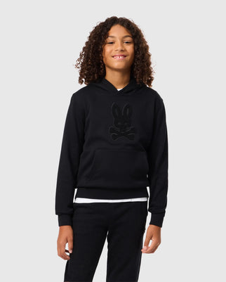 A person with curly hair is smiling at the camera while wearing the Psycho Bunny KIDS PACIFIC CHENILLE HOODIE - B0H930D200, against a plain gray background.