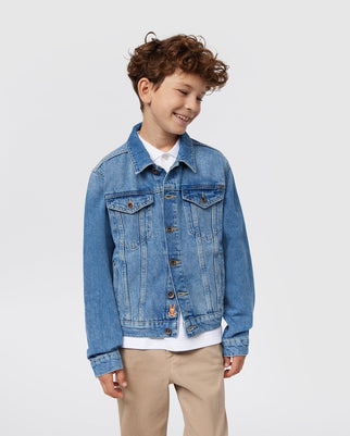 A smiling young boy with curly brown hair wears a blue KIDS PATHE DENIM JACKET - B0J741X1CO by Psycho Bunny made from recycled cotton over a white shirt and beige pants. He stands against a plain white background.