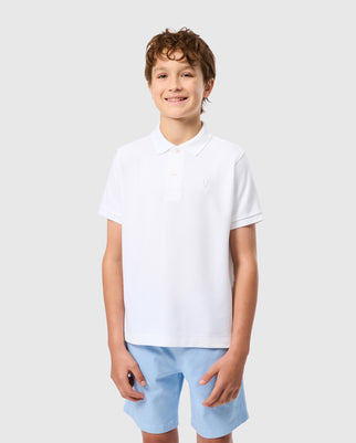 A smiling boy with curly hair is wearing a white Kids Classic Pique Polo Shirt (B0K021AR00) by Psycho Bunny, featuring an embroidered chest Bunny logo on the left side, paired with light blue shorts. He is standing against a plain light gray background.