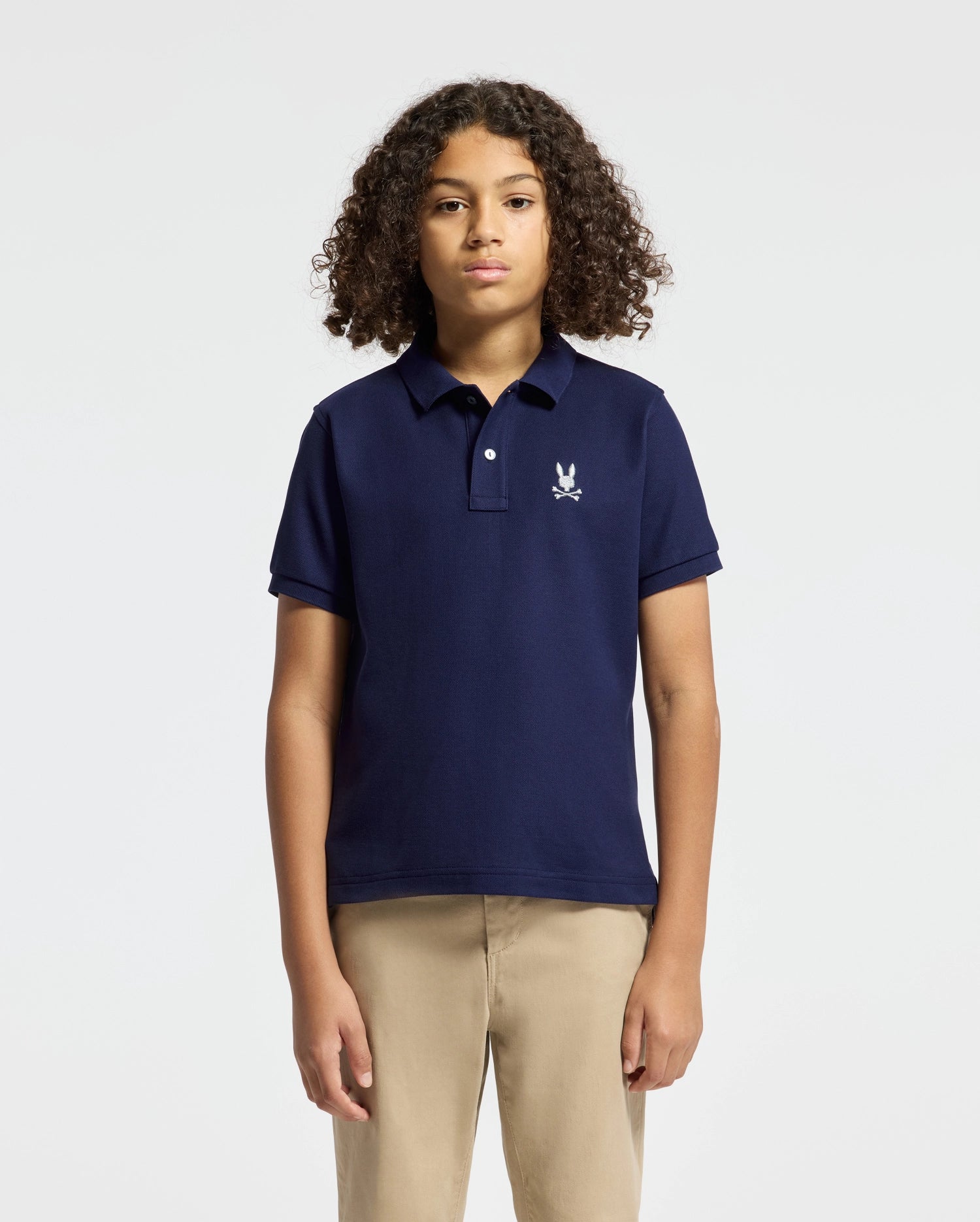 A young person with curly hair stands against a plain background, wearing a Psycho Bunny KIDS ALEXANDER PIQUE POLO in navy. The embroidered logo on the left chest of the pima cotton polo adds sophistication, paired effortlessly with tan pants.