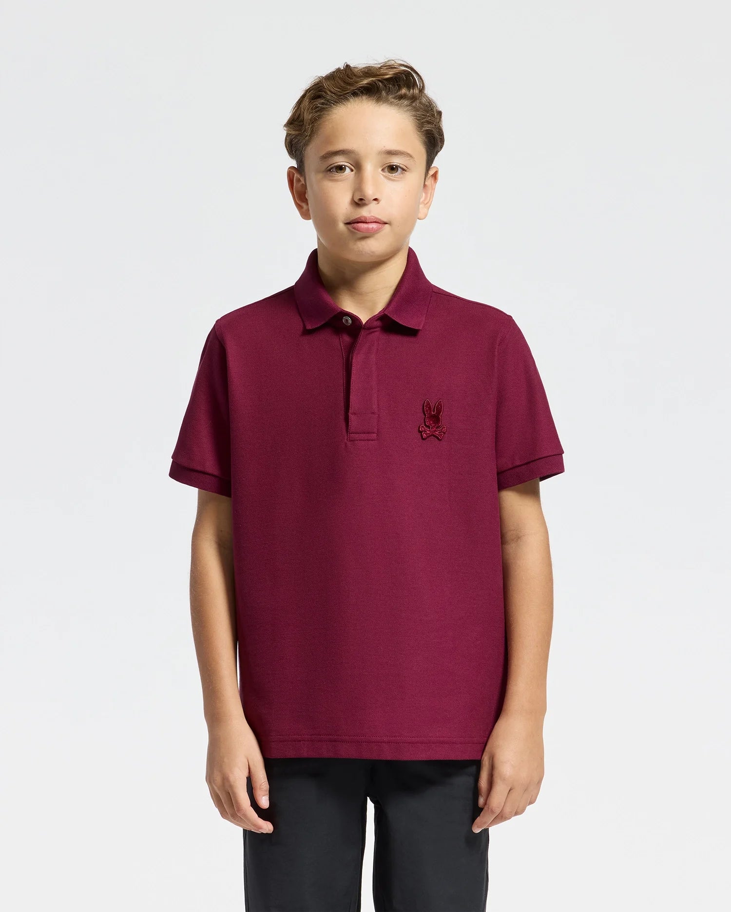 A young boy stands against a plain background, wearing a stylish Psycho Bunny Kids Brandson Pique Polo (B0K108E200) in burgundy with a small embroidered logo on the chest and black pants. His hair is short and styled neatly.