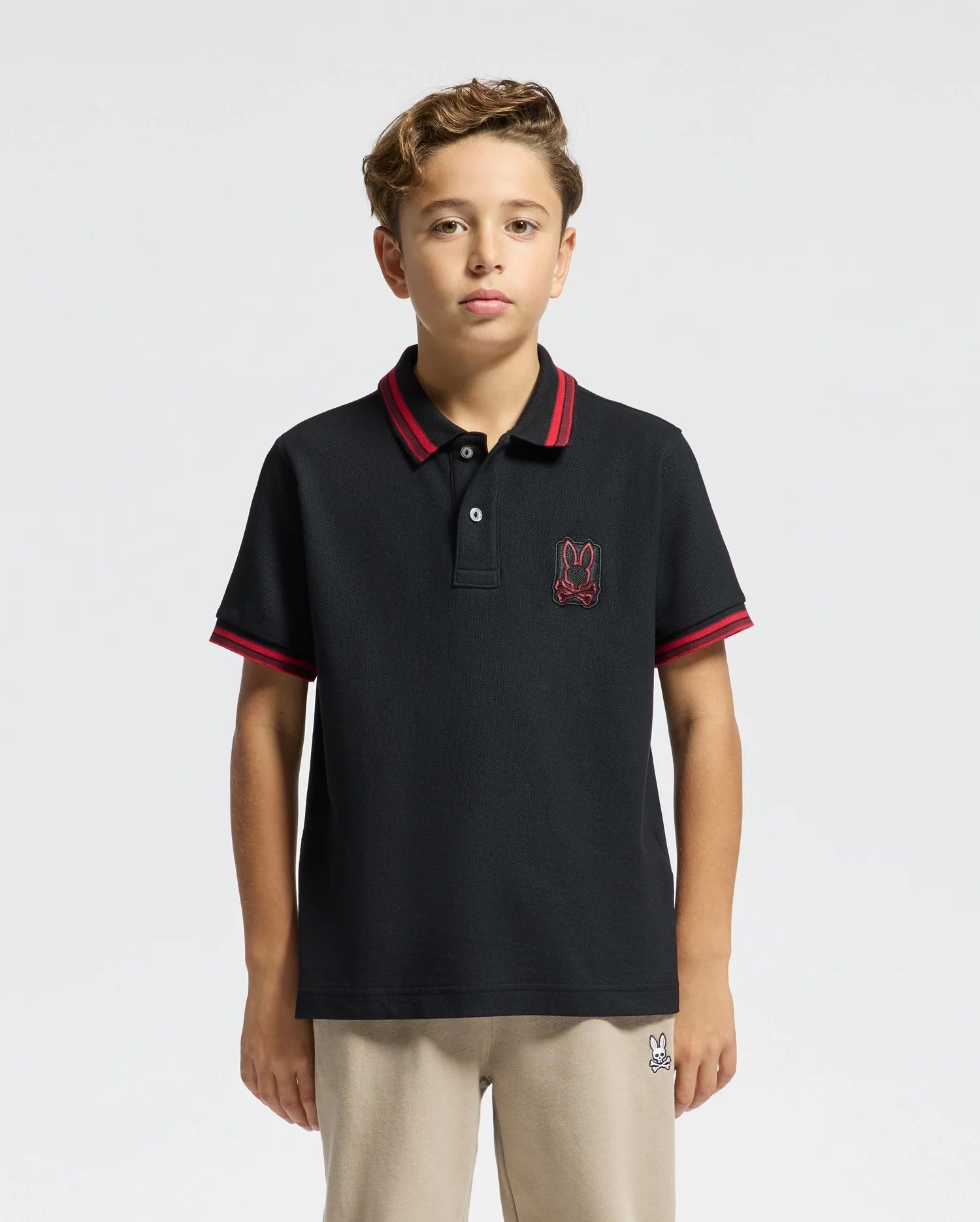 A young boy with curly hair is wearing the Psycho Bunny KIDS PIERCE PIQUE POLO - B0K201E200, which is crafted from Pima cotton and features red trim and an embroidered bunny logo. He pairs it with light-colored pants against a plain, light background.