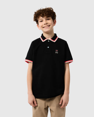 A smiling boy with curly hair is wearing a KIDS KINGSBURY PIQUE POLO SHIRT - B0K235B200 by Psycho Bunny in black, featuring red and white striped accents on the collar and sleeves. There is a small embroidered logo of a bunny on the left side of the Pima cotton shirt. He is standing against a plain light gray background.