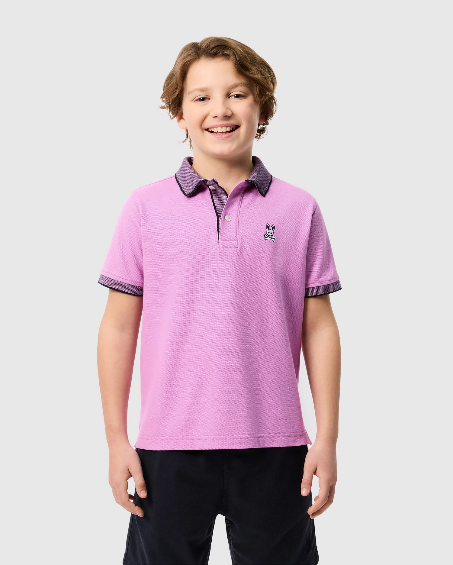 A smiling young boy with wavy hair poses against a plain, light gray background. He is wearing a light purple Psycho Bunny Southport pique polo shirt (B0K263B200) with a dark collar and sleeve edges, made from soft Pima cotton and featuring a small embroidered dog logo on the chest. Additionally, he is dressed in dark shorts.