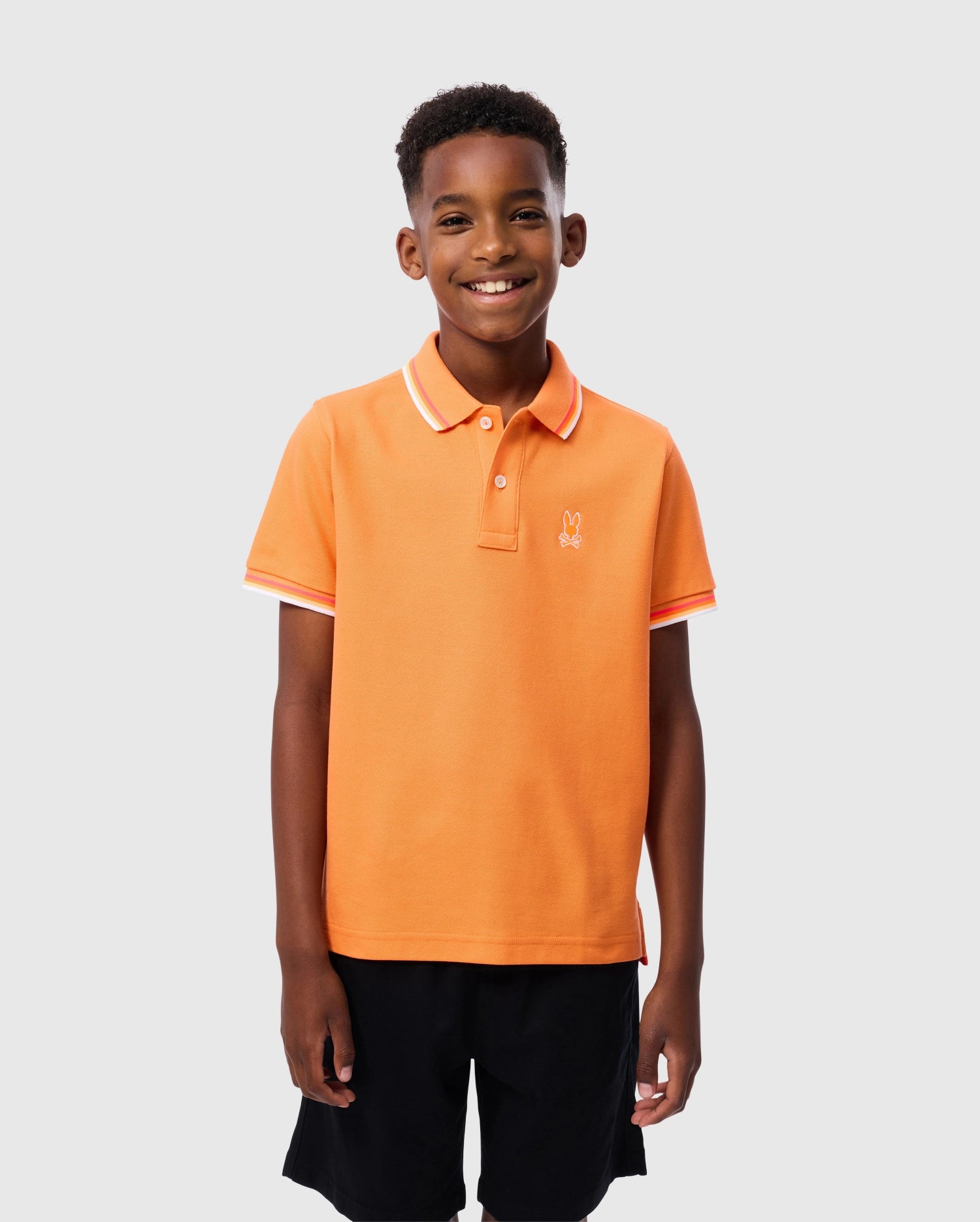 A young boy with short, curly hair is smiling at the camera. He is wearing an orange Kids Granbury Piqué Polo by Psycho Bunny, which features white accents on the rib knit collar and sleeve edges, paired with black shorts. The polo shirt, made of diamond-knit Pima cotton piqué, displays a small rabbit logo on the chest. The background is plain light gray.