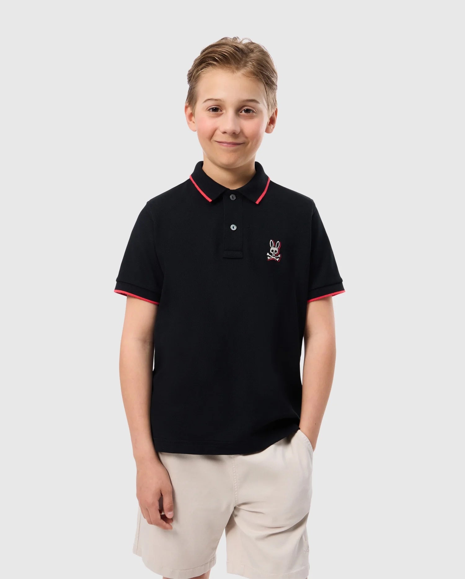 A smiling young boy with short light brown hair is wearing a Psycho Bunny Kids Kayden Pique Polo (B0K675C200) made of Pima cotton piqué, featuring contrasting stripes on the collar and sleeves. The shirt has a small embroidered logo of a bunny skull and crossbones on the left chest. He is also wearing beige shorts and has his hands in his pockets.