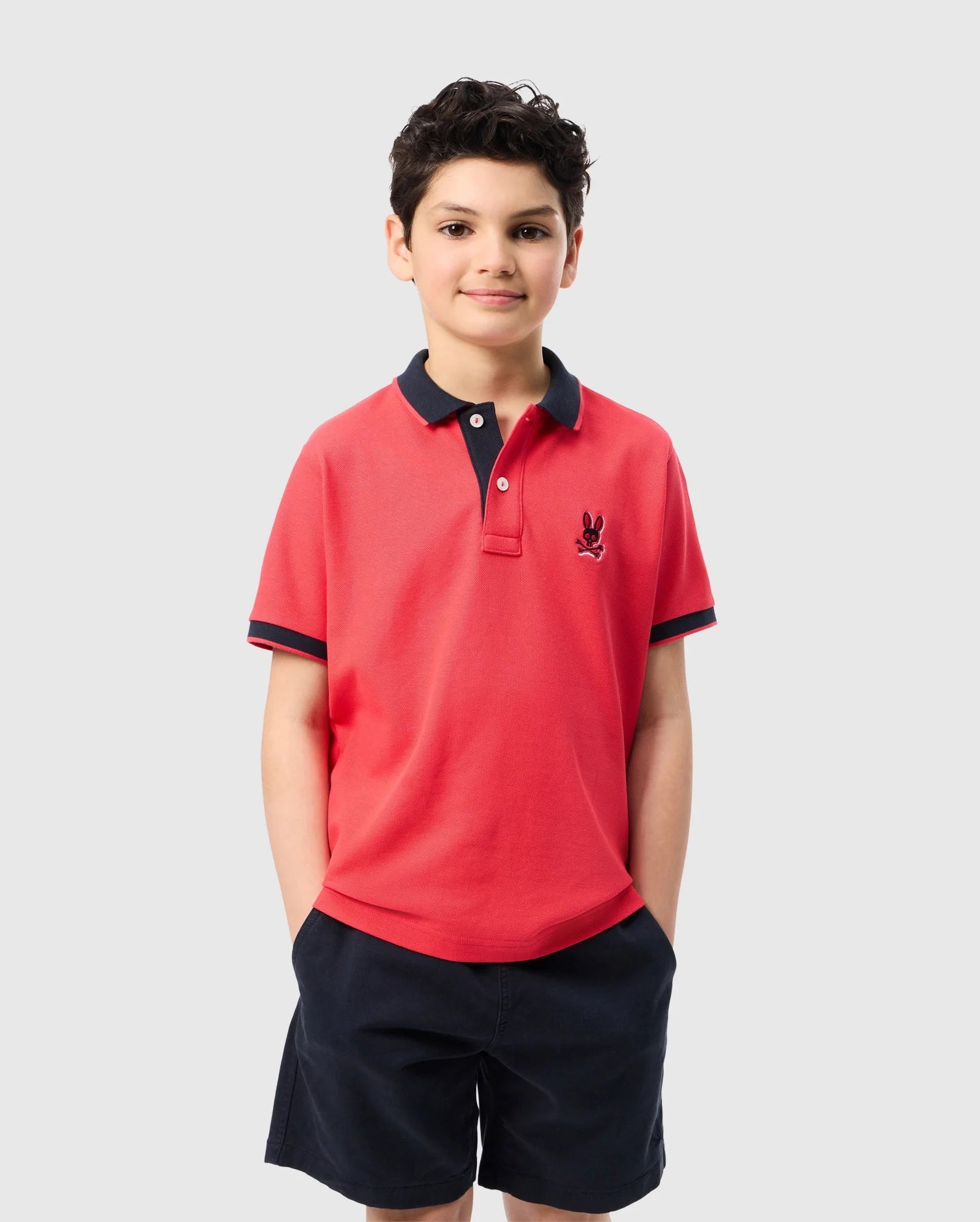 A young boy is standing against a plain white background. He is wearing a red Kids Kayden Pique Polo (product code B0K675C200) by Psycho Bunny, which features a navy blue collar and cuffs, along with a small logo on the left side of the chest. Paired with navy blue shorts, the polo is crafted from Pima cotton piqué to ensure comfort. He has short dark hair and a calm expression.