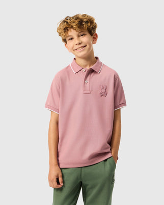 A cheerful child with curly hair is wearing the KIDS KINGSTON PIQUE POLO SHIRT - B0K764D200 by Psycho Bunny, made of soft Pima cotton in pink and featuring an embroidered Bunny patch on the chest. The child is paired with green pants and stands against a plain light gray background.