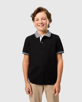 A smiling young boy with curly hair is standing against a plain background. He is wearing the Psycho Bunny Kids Malton Pique Polo Shirt (B0K950D200), which is made from Pima cotton and features white and gray accents on the collar and sleeves, paired with beige pants. His hands are relaxed by his sides.