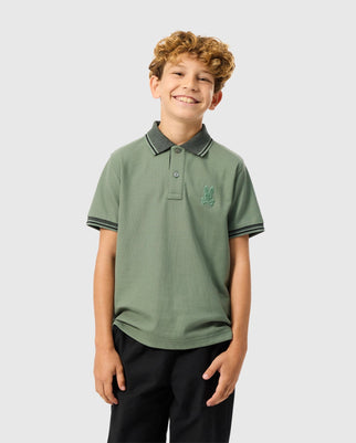 A smiling young boy with curly hair stands against a plain background. He is wearing the Psycho Bunny KIDS MALTON PIQUE POLO SHIRT - B0K950D200, an airy and breathable light green polo shirt made from 100% pima cotton. The shirt features a dark collar and sleeve trim and is tucked into black pants. An embroidered logo adorns the left side of his chest. His hands are resting by his sides.