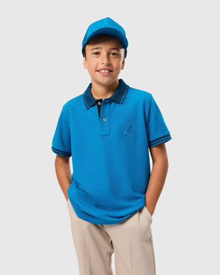 A young boy smiles while wearing a blue KIDS MALTON PIQUE POLO SHIRT - B0K950D200 and a matching blue cap. The Psycho Bunny polo, made from Pima cotton, features short sleeves, a collar, and an embroidered Psycho Bunny logo on the chest. He has his hands in the pockets of his beige pants and stands against a plain, light gray background.