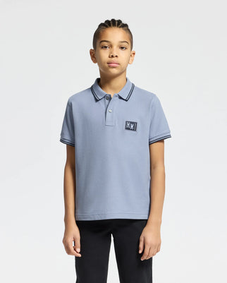 A young person with braided hair is wearing Psycho Bunny's KIDS MONROE PIQUE POLO in a regular fit, crafted from light blue Pima cotton and featuring intricate embroidered detailing on the chest. They have paired it with black pants and are standing against a plain white background.