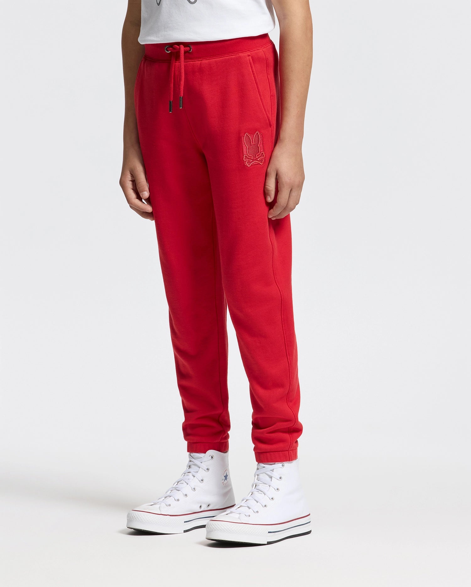 Someone is wearing Psycho Bunny KIDS PIERCE ESSENTIAL FRENCH TERRY SWEATPANT, featuring an embroidered logo on the left side. They pair it with white high-top sneakers and a white top partially visible against a plain white background.