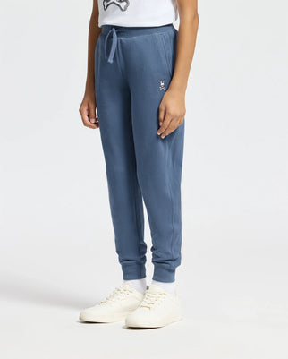 A person wearing Psycho Bunny's KIDS LEVY ESSENTIAL FLEECE SWEATPANT in blue, crafted from 100% cotton, and a white t-shirt stands against a plain background. The casual look is complemented by a small embroidered logo on the left thigh of the jogger pants, paired with white sneakers and socks.