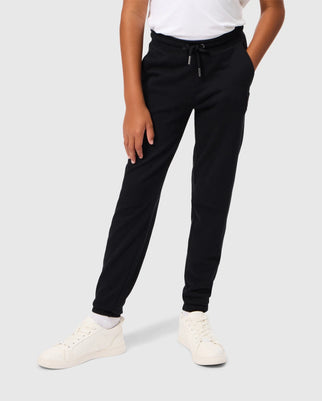 A person dressed in a white T-shirt, the Psycho Bunny KIDS PACIFIC CHENILLE JOGGER - B0P972D200 in black with a stylish chenille logo patch, and white sneakers is standing with one hand in their pocket against a plain light gray background.