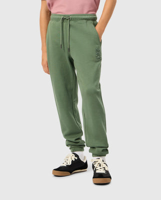 A person wearing a pink shirt and green Psycho Bunny KIDS PACIFIC CHENILLE JOGGER (B0P972D200) featuring a chenille logo patch on the upper left leg. These casual bottoms come with a drawstring waist and are styled with black sneakers that have white laces and a thick brown sole.