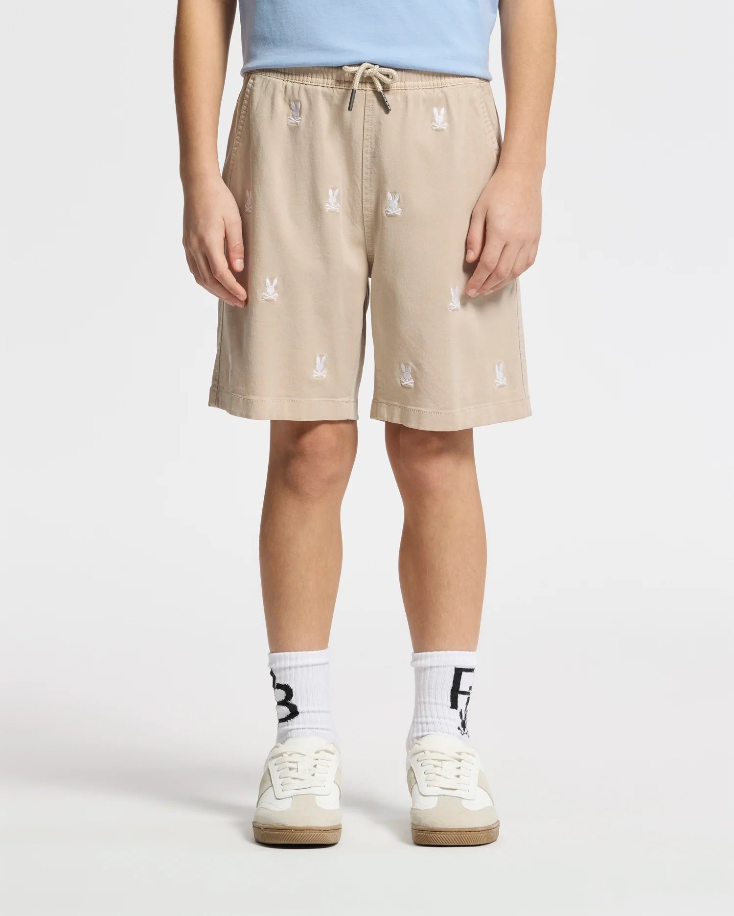 A person wears Psycho Bunny's Kids All Over Willis Stretch Tencel Shorts with embroidered bunnies, a light blue shirt tucked in, and white socks with black letters. Their white sneakers have beige soles. The scene is set against a plain white background, with hands resting casually by the sides.