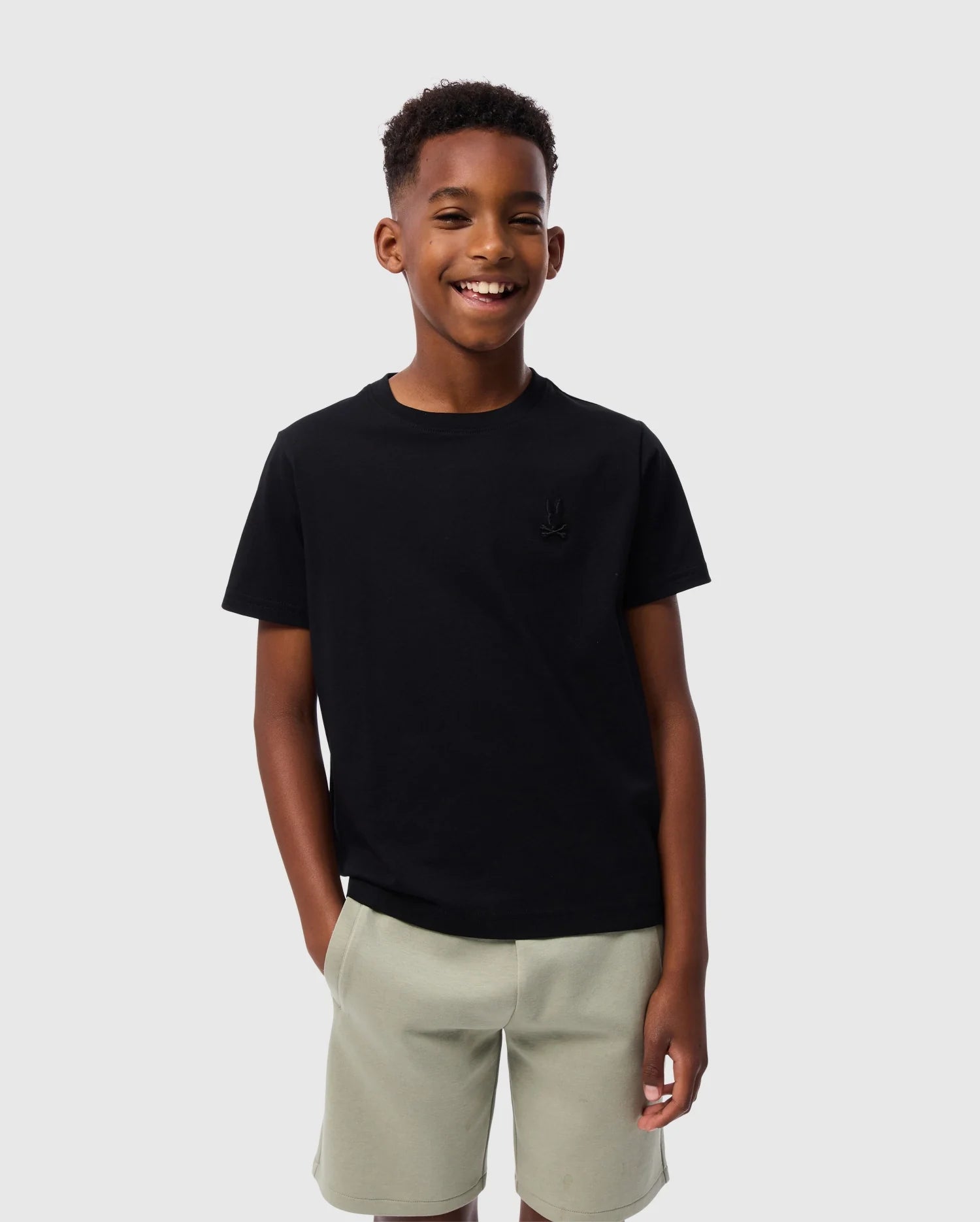 A smiling young boy is standing against a plain light gray background. He is wearing a black KIDS EMBROIDERED CLASSIC TEE - B0U020AR00 by Psycho Bunny and light gray shorts, with his hands casually resting in his pockets. His short curly hair adds to the charm of his relaxed look.