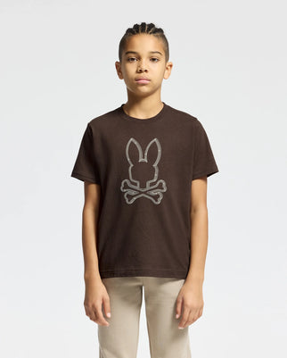 A young person with braided hair stands wearing the KIDS MONARC FRONT BUNNY GRAPHIC TEE - B0U211E200 from Psycho Bunny, a brown tee crafted from soft Pima cotton and showcasing a playful bunny and crossbones design. They pair it with light-colored pants against a plain white background for a minimalist look.