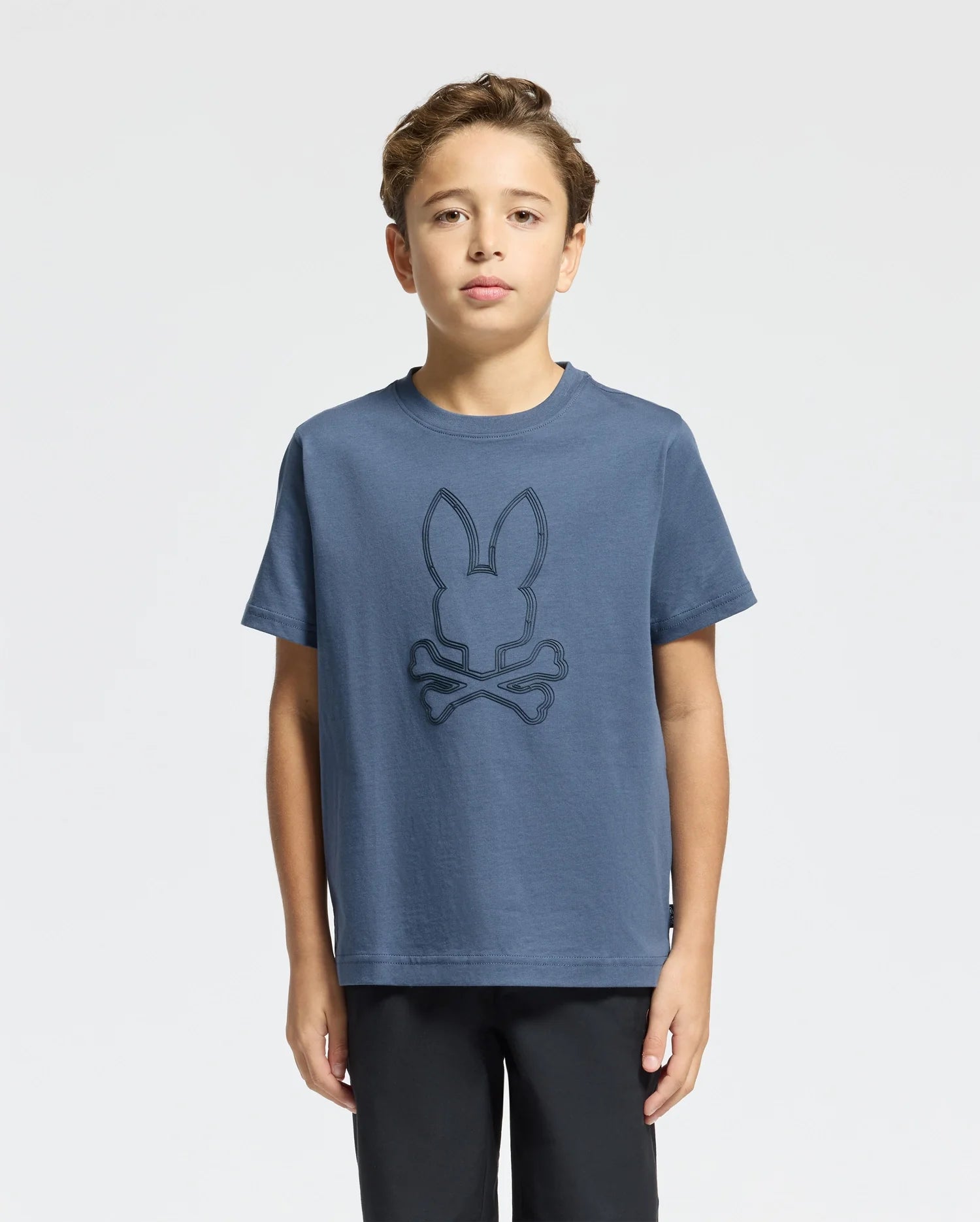 A young boy wearing the Psycho Bunny Kids Monarc Front Bunny Graphic Tee (B0U211E200), crafted from soft Pima cotton and showcasing a raised-printed bunny head with crossed bones design, stands against a plain background. He has short brown hair and is looking directly at the camera.