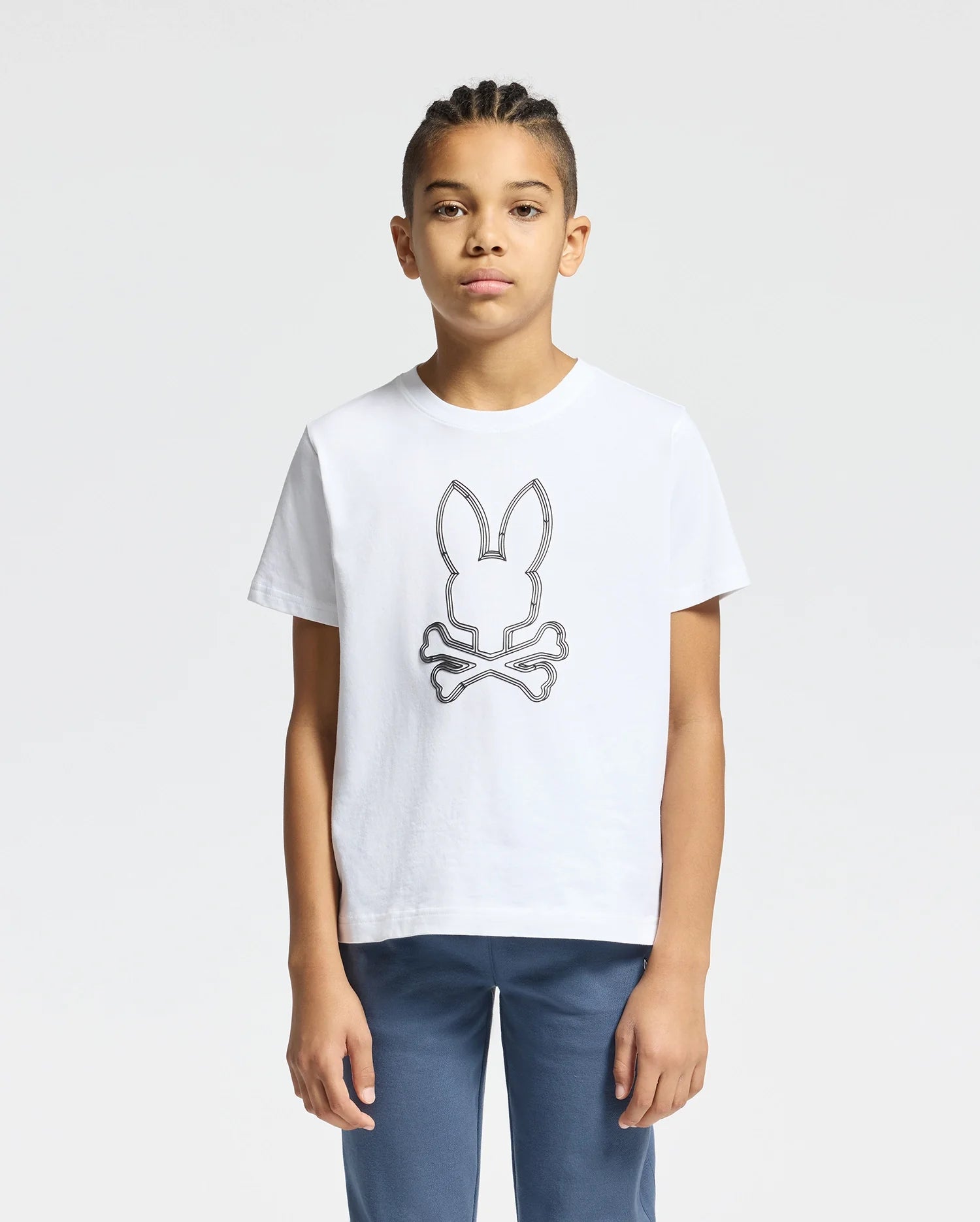 A young individual stands against a plain backdrop, wearing the KIDS MONARC FRONT BUNNY GRAPHIC TEE - B0U211E200 by Psycho Bunny, crafted from soft Pima cotton. This white tee showcases a striking design of bunny ears and crossbones. Paired with braided hair and blue pants, they effortlessly combine comfort with style.