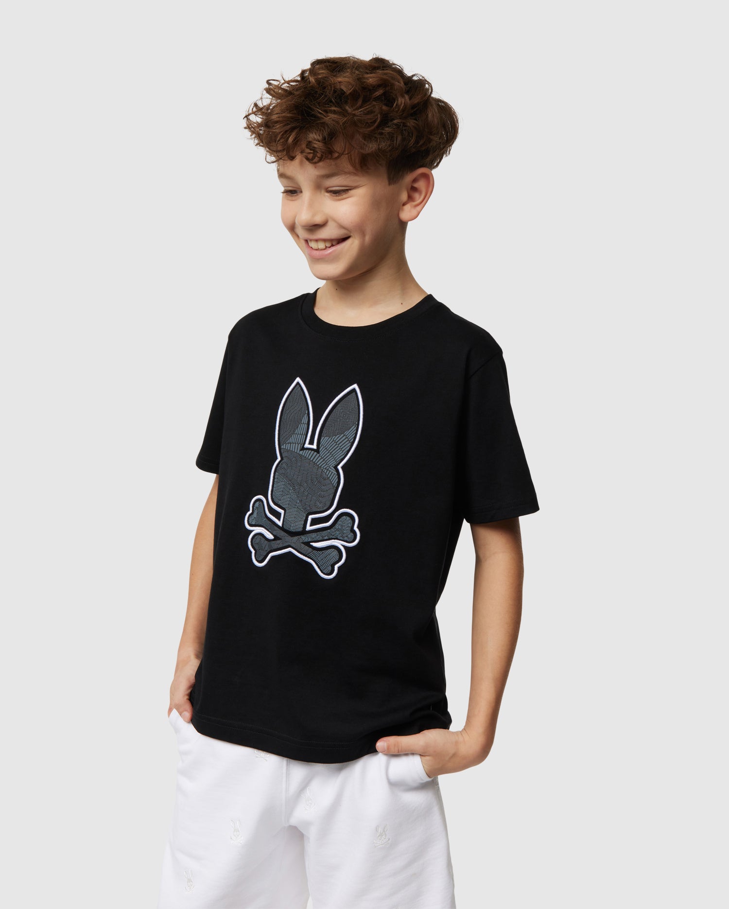 A cheerful boy with curly hair stands against a plain background, wearing a black Psycho Bunny KIDS LENOX EMBROIDERED GRAPHIC TEE - B0U405B200 featuring a rabbit skull and crossbones design. He is also wearing white pants made of soft Pima cotton, with one hand in his pocket while smiling.