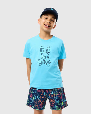 A smiling boy in a Psycho Bunny KIDS SHELDON GRAPHIC TEE - B0U569C200, adorned with a bunny skull and crossbones design, is matched with vibrant shorts featuring a similar pattern. He sports a dark cap against a light gray background. The outfit's playful style is further highlighted by the embroidered logo detail.