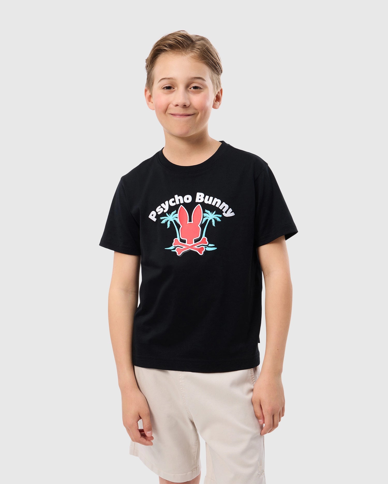 A smiling child with short light-brown hair is wearing the KIDS BOSTON GRAPHIC TEE - B0U573C200, a high-quality black cotton T-shirt from Psycho Bunny featuring a colorful logo of a bunny and crossed bones. The child pairs it with white shorts and stands against a plain white background.