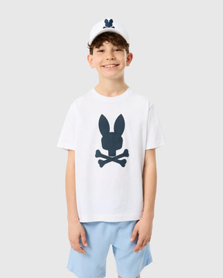 A young boy stands smiling, dressed in the Psycho Bunny KIDS HOUSTON GRAPHIC TEE - B0U607C200 made of Pima cotton, featuring a blue rabbit skull and crossbones design. He pairs it with light blue shorts and a white cap adorned with the matching blue rabbit logo. The background is plain gray.