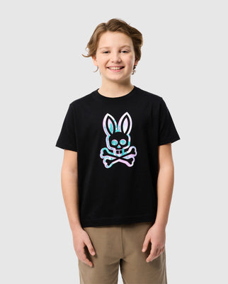 A young boy with wavy hair smiles while wearing a black KIDS LEONARD GRAPHIC TEE - B0U609C200 from Psycho Bunny, featuring a colorful bunny print skull and crossbones graphic. He is also wearing beige pants. The background is plain and light gray.